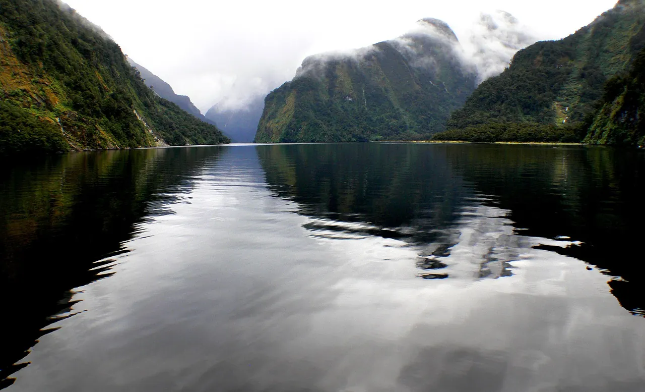 Doubtful Sound with forested walls reflected in flat water and cloud on the ridges